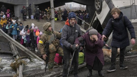 Muž pomáha dôchodkyni prejsť po provizórnom chodníku po moste zničenom ruským leteckým náletom v ukrajinskom meste Irpiň.