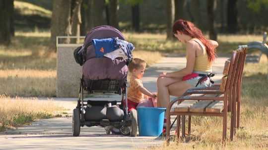 Matka sedí na lavičke v parku a dieťa sa vozí na odráždle.na