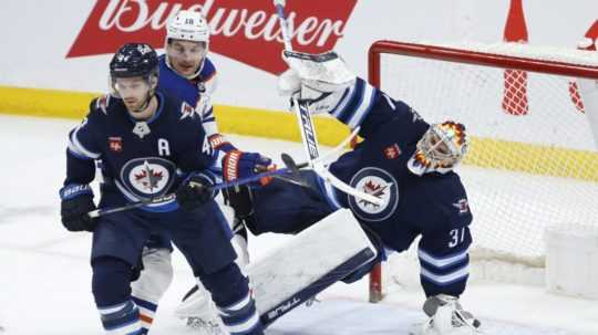 Brankár Winnipegu Jets Connor Hellebuyck (37) padá pri Zachovi Hymanovi (18) z Edmontonu Oilers, Josh Morrissey (44) bráni Jets počas tretej tretiny hokejového zápasu NHL vo Winnipegu.