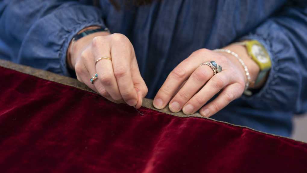 Členka britskej Kráľovskej školy vyšívania obšíva červenú korunovačnú róbu zlatým lemom.