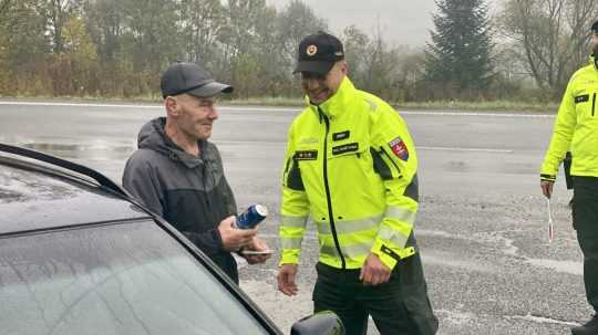 Na sn´mke stojí vodič s policajným príslušníkom pri aute držiac nealkoholické pivo.