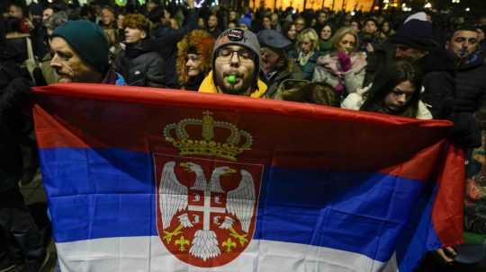 Muž píska a drží srbskú vlajku pred budovou volebnej komisie počas protestu opozície v Belehrade.