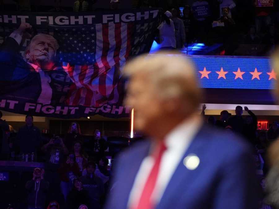 V popredí Donald Trump a jeho žena Melania, v pozadí veľká vlajka s portrétom psotreleného Trumpa.