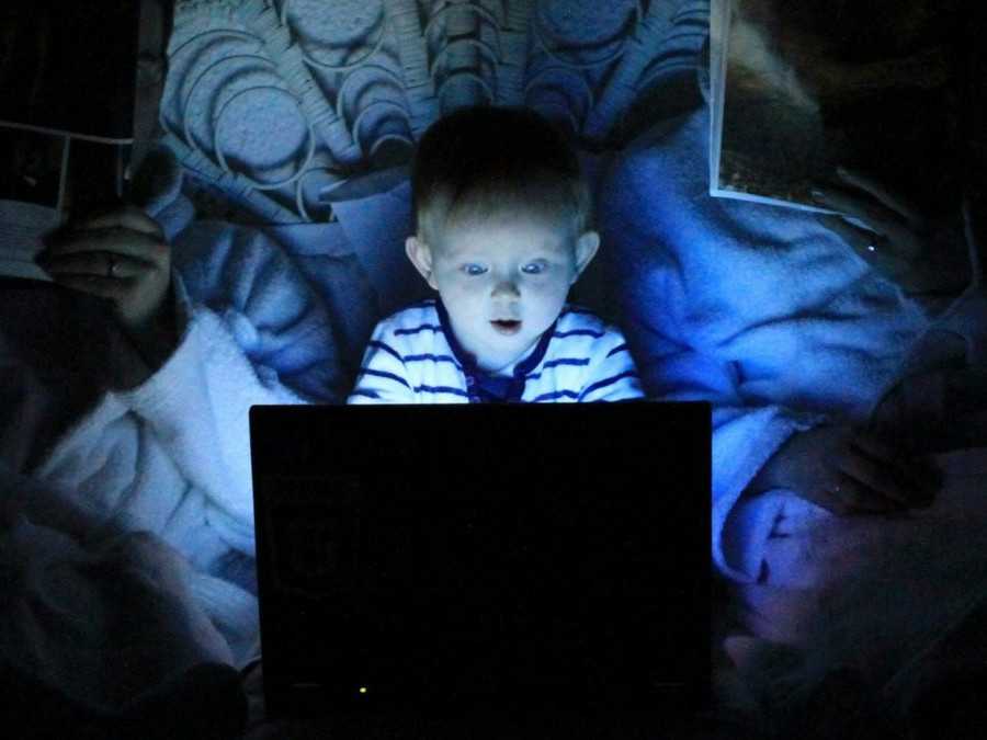 Malé dieťa v posteli pozerá na jasnú obrazovku laptopu. Rodičia po jeho boku majú zakryté tváre novinami.