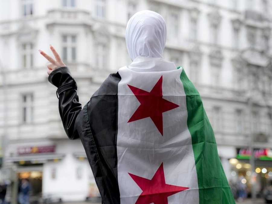 Sýrske dievča máva na ulici v Berlíne znakom víťazstva, pričom má na sebe vlajku sýrskej opozície.