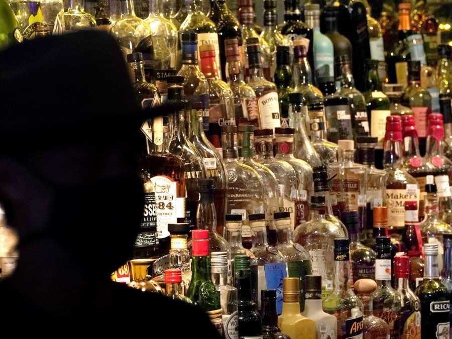 Na snímke v popredí silueta muža s klobúkom, v pozadí fľaše s alkoholom za barom.