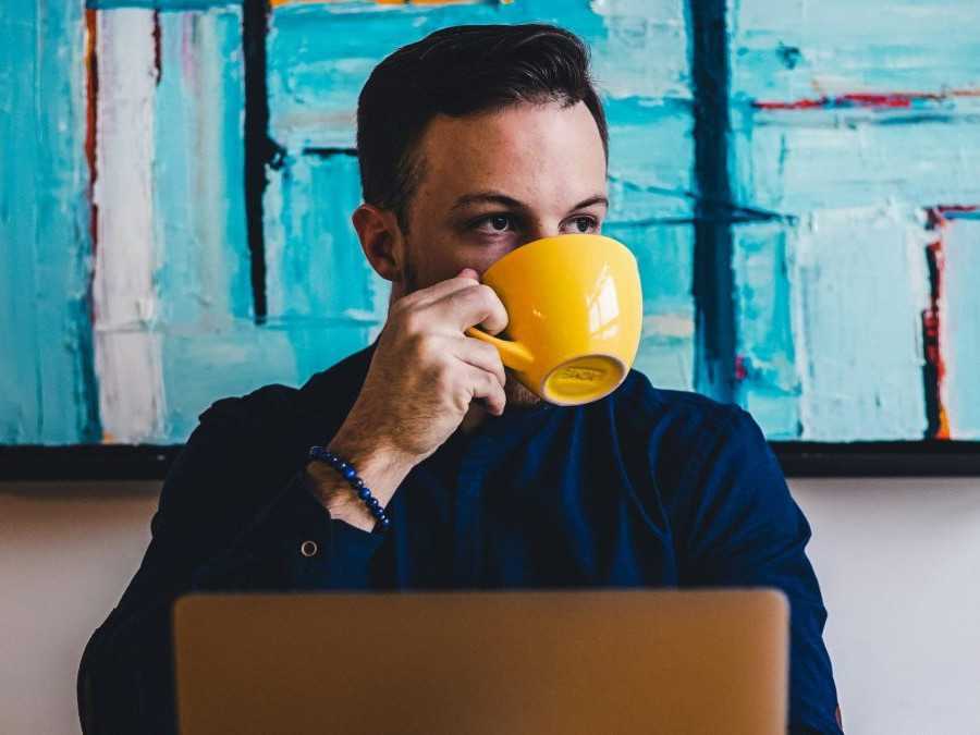 Na snímke je muž, pije kávu za stolom, na ktorom je položený laptop.