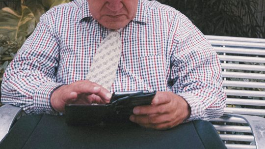 Na snímke senior v košeli s kravatou pracuje so smartfónom.