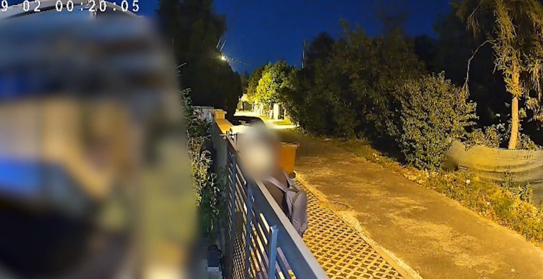 Kamera zachytáva muža, ako nazerá za plot rodinného domu.