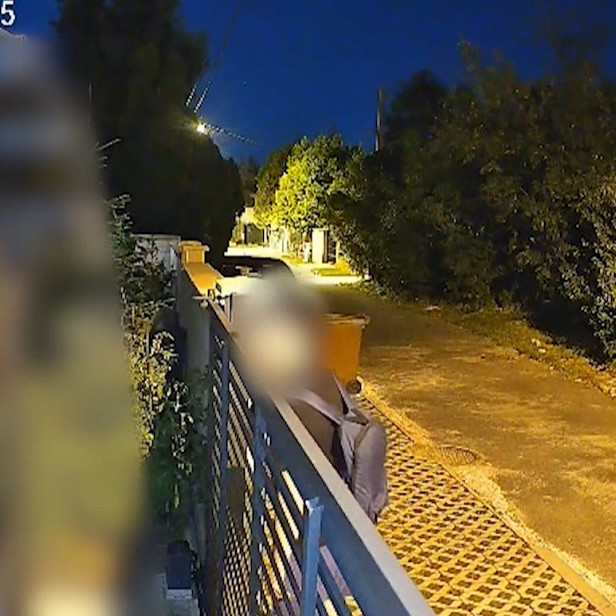 Kamera zachytáva muža, ako nazerá za plot rodinného domu.