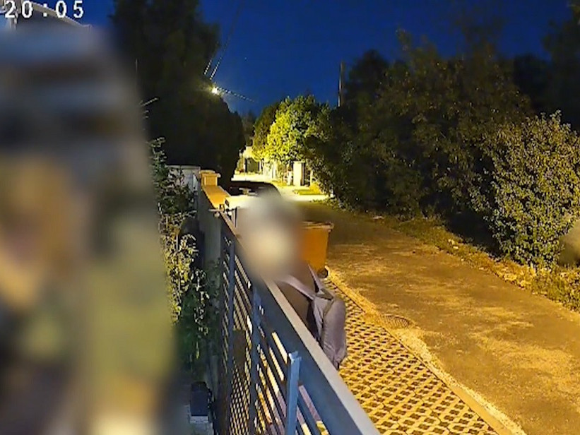 Kamera zachytáva muža, ako nazerá za plot rodinného domu.