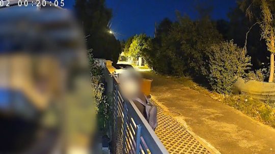 Kamera zachytáva muža, ako nazerá za plot rodinného domu.