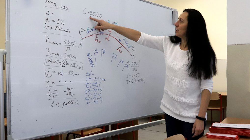 Na snímke je učiteľka, ktorá na tabuli vysvetľuje učivo z matematiky.