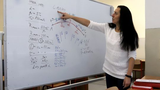 Na snímke je učiteľka, ktorá na tabuli vysvetľuje učivo z matematiky.