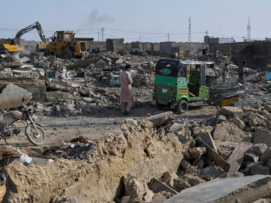 Na snímke ľudia prechádzajú cez trosky, zatiaľ čo hydraulická lopata búra budovu počas operácie proti nelegálnemu osídleniu afganských utečencov.