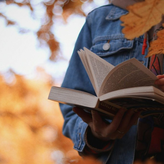 Osoba číta knihu, v popredí a v pozadí listy stromu.