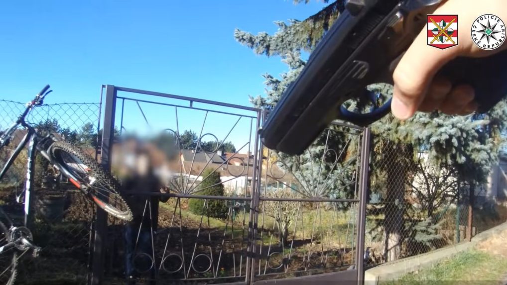 Na snímke policajt mieri na recidivistu za plotom zbraňou.