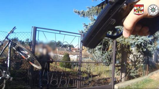 Na snímke policajt mieri na recidivistu za plotom zbraňou.