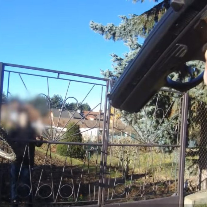 Na snímke policajt mieri na recidivistu za plotom zbraňou.