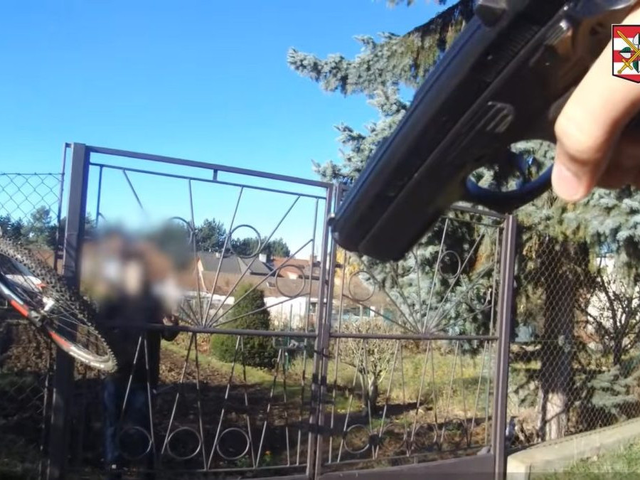 Na snímke policajt mieri na recidivistu za plotom zbraňou.