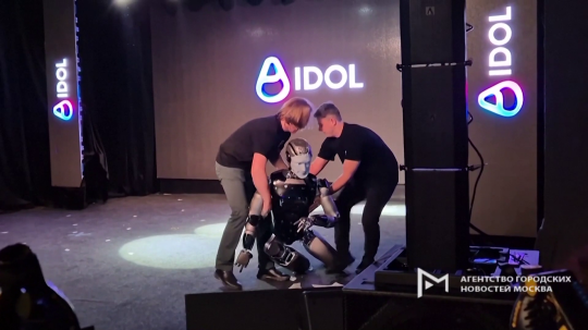 Na snímke je ruský robot poháňaný AI, ktorého drží z každej strany dvojica mužov po tom, ako na pódiu spadol.