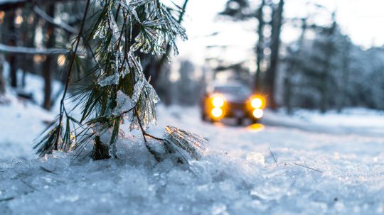 Na snímke sa auto pohybuje po zasneženej ceste, v popredí je detail na zasneženú vetvičku ihličnatého stromu.