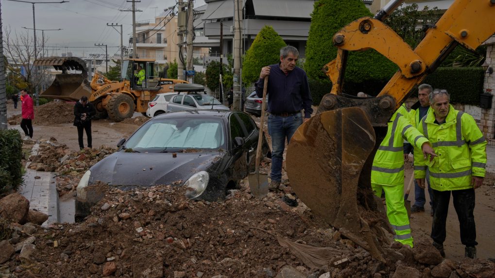 Pracovníci mestských služieb a obyvatelia odstraňujú blato a trosky z ulice vedľa poškodeného auta po povodniach spôsobených silnými dažďami.