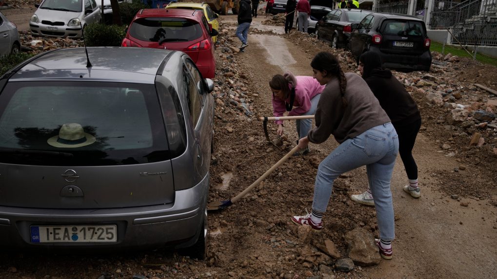 Pracovníci mestských služieb a obyvatelia odstraňujú blato a trosky z ulice vedľa poškodeného auta po povodniach spôsobených silnými dažďami.